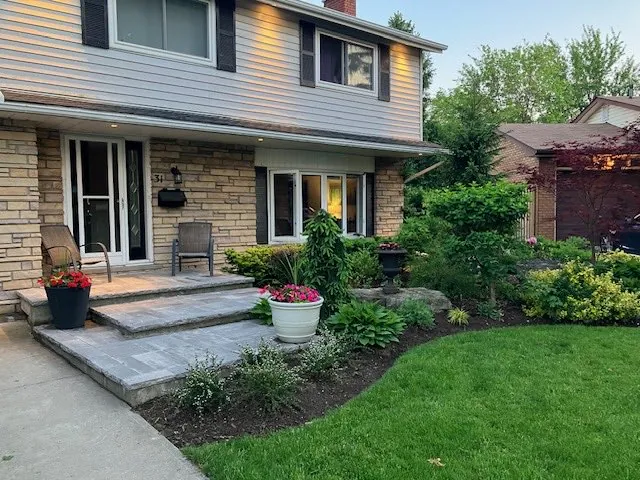 Freshly landscaped front entrance at dusk with layered plantings, potted flowers and a paver step, by Walnut Creek Landscaping in Hamilton