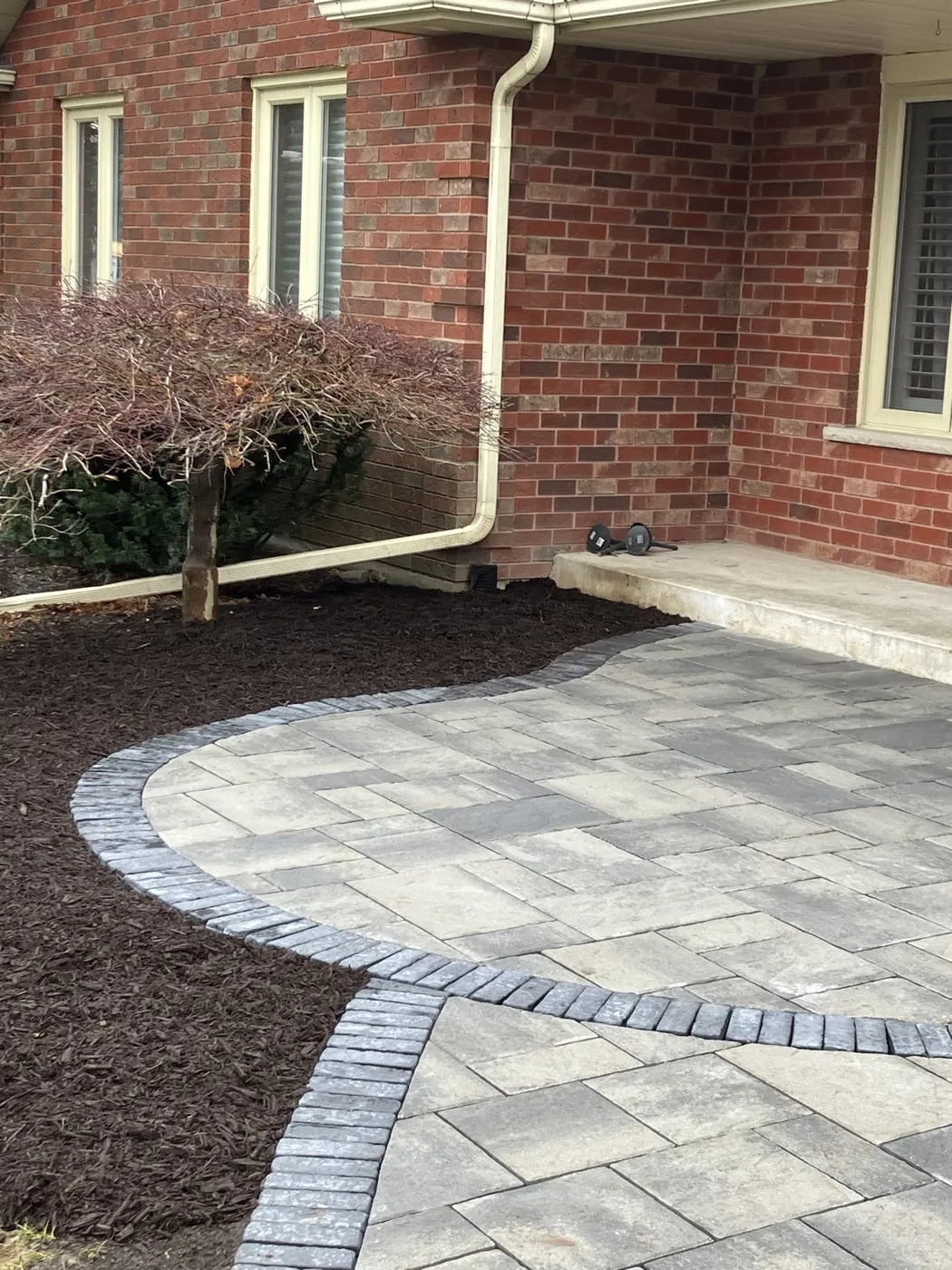 Grey paver walkway with a charcoal border curving to a home's entrance with spring plantings in Hamilton