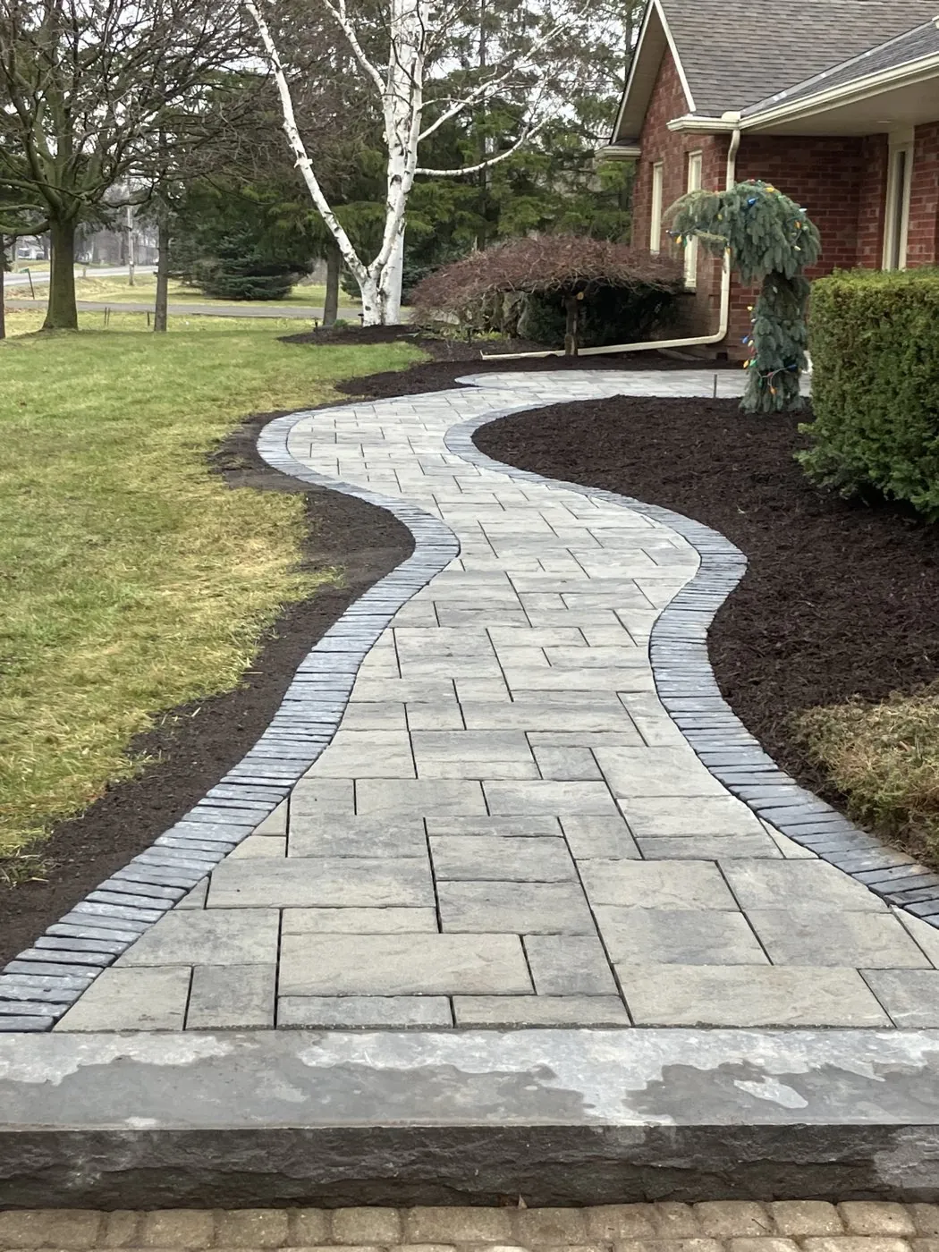 Winding grey paver walkway with a soldier-course border curving through mulched garden beds to a Hamilton home